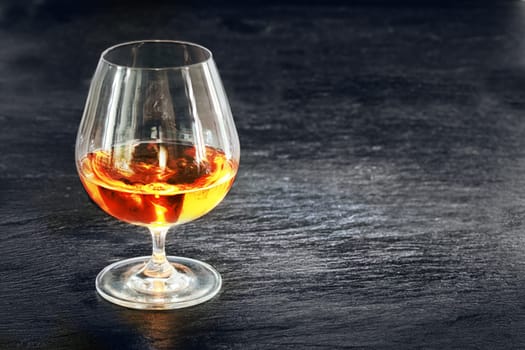 Elegant snifter with glowing golden cognac standing on a rustic dark bar counter with copyspace