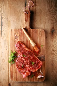 Seasoning a t-bone steak for grilling with BBQ or chili based spice using a small brush on a wooden board with fresh rosemary, overhead view