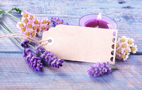 Spa or wellness background with a blank decorative gift tag surrounded by dainty fresh flowers and lavender with a burning aromatherapy candle with essential oil on rustic lilac colored wooden boards