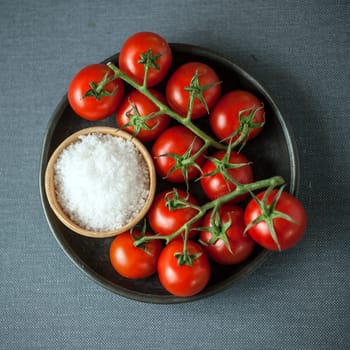 Overhead view of a bunch of ripe red cherry tomatoes on the vine and a bowl of coarse natural sea salt for cooking ingredients on a grey cloth background in square format