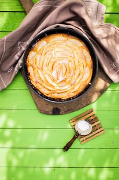 Fresh home baked apple pie in an oven dish with a small strainer with powdered sugar to sprinkle on top on a green garden table in dappled sunlight viewed from overhead