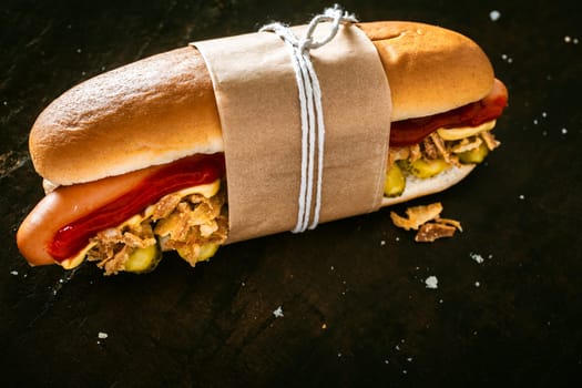 Delicious hot dog with a smoked Vienna sausage and salad ingredients with tomato sauce and mustard tied in a brown wrapper against a dark background