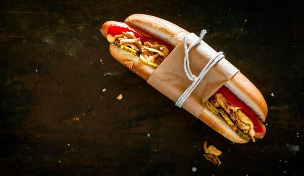 Hot dog with all the trimmings with a smoked frankfurter, pickles, salad and mustard tied in a brown paper wrapper against a black background with copyspace