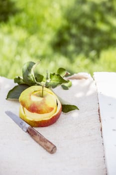 Peeled fresh healthy apple with a paring knife and leaves on a white cloth on a painted wooden rustic table outdoors in a lush summer garden