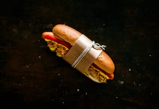 Takeaway hot dog with a smoked Wiener sausage and all the trimmings on a fresh bun tied with brown paper and string on a black background with copyspace