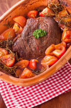 Pot roast with tomatoes, herbs and a portion of browned braised beef in a terracotta casserole dish simmered in a rich gravy or sauce
