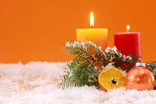 Warm orange themed Christmas background with two burning candles with a pine branch , bauble and slice of dried orange nestling in snow against an orange background with copyspace for your greeting