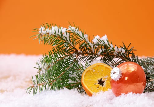 Orange themed Christmas background with a shiny bauble and dried orange slice nestling in wintry snow with a pine branch against an orange background with copyspace