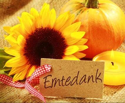 Colorful Thanksgiving background with a gift tag with the German word - Erntedank - with a vibrant yellow sunflower orange pumpkin and burning candle grouped on rustic hessian