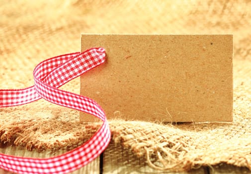 Blank brown tag with copyspace tied with a fresh red and white rustic ribbon on a piece of coarse woven hessian fabric for a natural country background