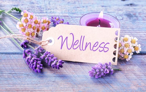 Pretty floral Wellness background with fresh scented lavender and dainty inflorescences around a label with the script - Wellness - leaning against a burning purple aromatherapy candle in a spa
