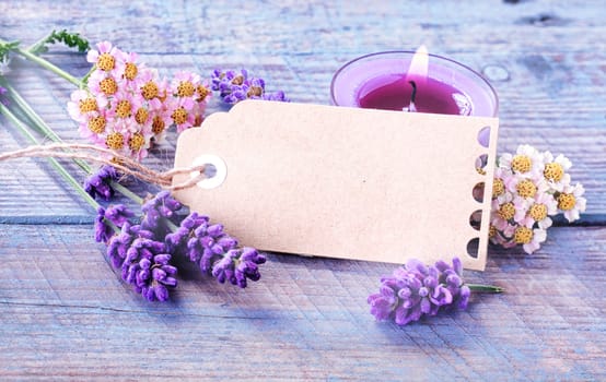 Spa, relaxation and wellness background with a blank gift tag or label with copyspace amongst fresh lavender and flowers with a burning candles for aromatherapy treatment on rustic blue wooden boards