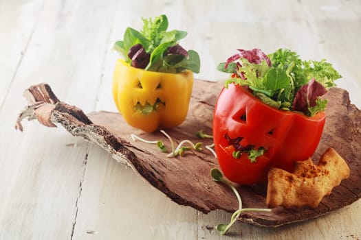 Colorful fresh Halloween bell pepper salads in individual servings for appetizers with carved jack-o-lantern faces stuffed with leafy green lettuce and herbs served on a bark platter with a bat cookie