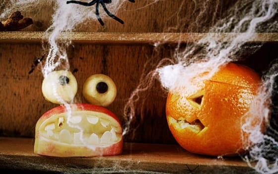 Fun spooky homemade Halloween food decorations with a carved apple mouth full of fearsome teeth topped with round dough eyes and a jack-o-lantern carved orange on a wooden shelf with spiders and webs