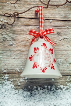 Decorative ceramic Christmas bell tied with a red and white checked ribbon hanging from rusty wire on a rustic background with snowflakes below