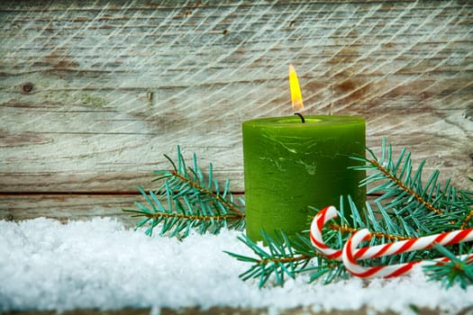 Christmas still life with a burning candle standing in winter snow with pine foliage and colorful red and white striped candy canes, wooden background with copyspace