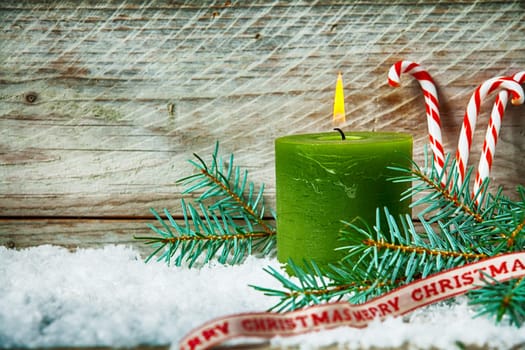 Lit Christmas candle amongst pine in winter snow with colorful red and white striped candy canes and a decorative ribbon with the message - Happy Christmas - wooden boards background with copyspace