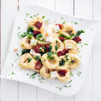 Overhead view of a plate of stuffed Italian tortellini pasta with feta cheese and pesto sprinkled with fresh herbs and pine nuts