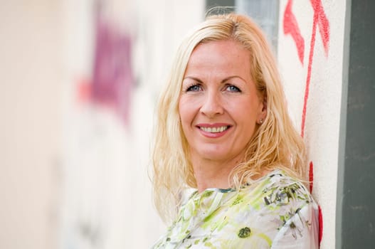 Side head portrait of a middle-aged blonde woman with her back standing on a painted wall and looking into the camera