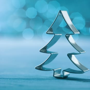Shiny silver cookie cutter Christmas tree decoration on a wintry cold blue background with a bokeh of twinkling party lights and copyspace , square format