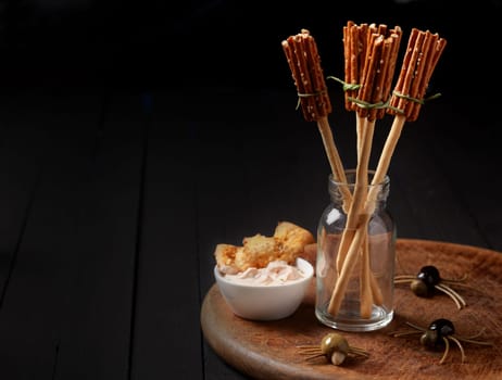 Witches broomsticks at a Halloween party made from bread sticks and pretzels standing in a glass jar on a table decorated with creepy spiders made from olives and spaghetti