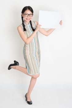 Portrait of excited Asian Chinese female hands holding a blank white card looking at camera, in retro revival style cheongsam, standing on plain background.