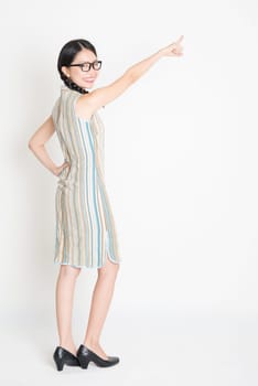 Portrait of rear view full length Asian Chinese girl pointing on blank copy space and looking at camera, in retro revival style cheongsam, standing on plain background.