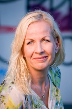 frontal head portrait of a middle-aged blonde woman in front of a painted wall friendly smiling at the camera