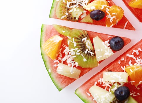 Close up overhead detail of a slice of tropical fruit watermelon pizza topped with kiwifruit, blueberries, orange, pineapple, and sprinkled with desiccated coconut, on white with copyspace