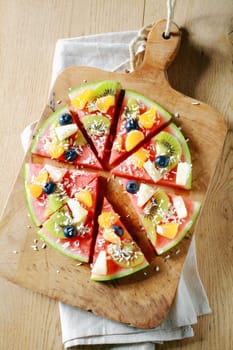 Overhead view of a colorful tropical fruit watermelon pizza topped with kiwifruit, blueberries, orange, pineapple, and sprinkled with desiccated coconut cut into segments on a rustic wooden board