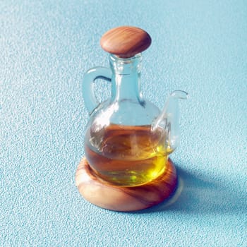 Clear glass decanter of golden virgin olive oil on a wooden stand for serving as a salad dressing or to use as an ingredient in cooking, on blue