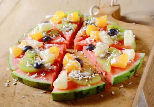 A tasty juicy fresh tropical fruit watermelon pizza topped with kiwifruit, blueberries, orange, pineapple, and topped with dried coconut, cut into segments on a wooden board for a nutritious dessert