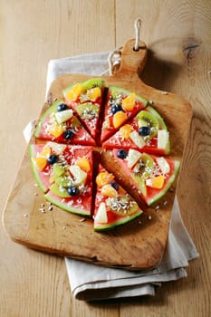 View from above of a sliced colorful tropical fruit watermelon pizza topped with kiwifruit, blueberries, orange, pineapple, and sprinkled with dried coconut on a wooden board for a fun party dessert