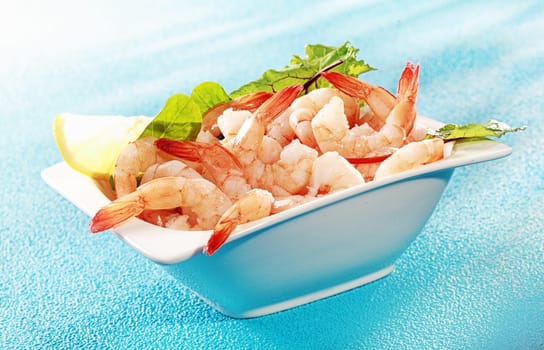Delicious seafood appetizer of grilled shrimp or pink prawns served in a dish with a slice of lemon and salad greens on a turquoise blue background