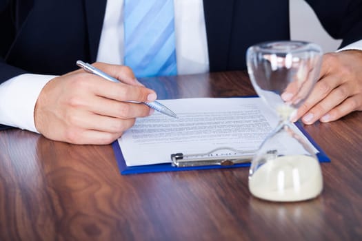 Close-up Of Businessman Filling Form With Hourglass On Desk