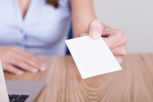 Young businesswoman sitting behind her laptop holds out her hand offering her card with focus to her face