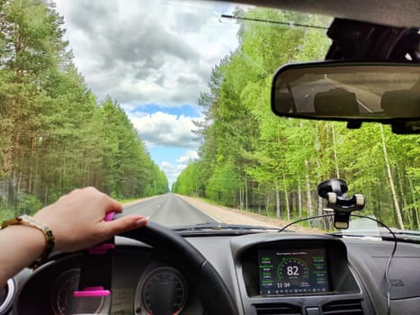 view through car windshield of natural landscape with road, green tree, white clouds. Hand of woman on steering wheel of car. Female traveler driving on trip or journey in spring or summer
