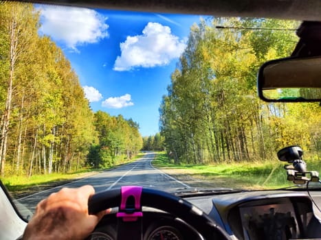 view from a car windshield of natural landscape with road, green trees and blue sky in summer or spring time. Hand of woman on steering wheel. Female traveler driving alone on trip or journey