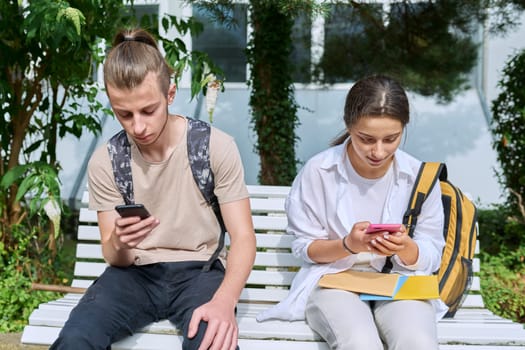Teenage high school students, guy and girl, sitting on bench with backpacks, near academic building, using smartphones. Adolescence, youth, education, lifestyle, technology concept