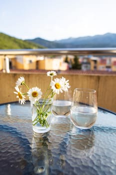 A bouquet of daisies in a glass vase with water stands on a glass table along with a glass of water