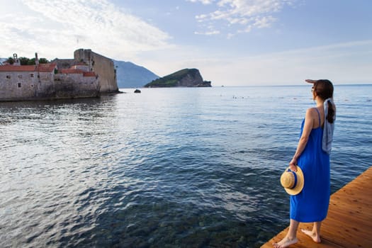 A girl in a blue dress with a straw hat stands on a wooden pier near the sea. In the background is the old medieval town in Montenegro in Budva