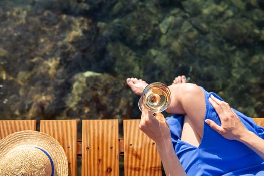 A girl holds a glass of wine in her hand while sitting on a wooden pier near the Adriatic Sea. Straw hat, vacation, travel. View from above