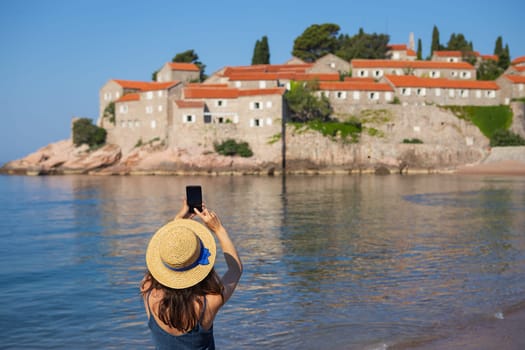 Sveti Stefan Island, Montenegro July 5, 2021: Adriatic Sea. A girl in a straw hat stands and photographs the island of St. Stephen from the beach