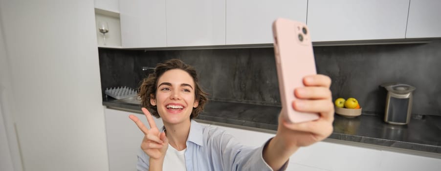 Happy, beautiful brunette girl takes selfie at home, poses for photo with smartphone.