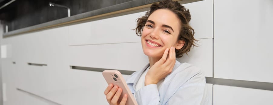Close up portrait of happy woman with a phone at home, she touches her face and smiles lightly, uses mobile application. Technology and people concept.