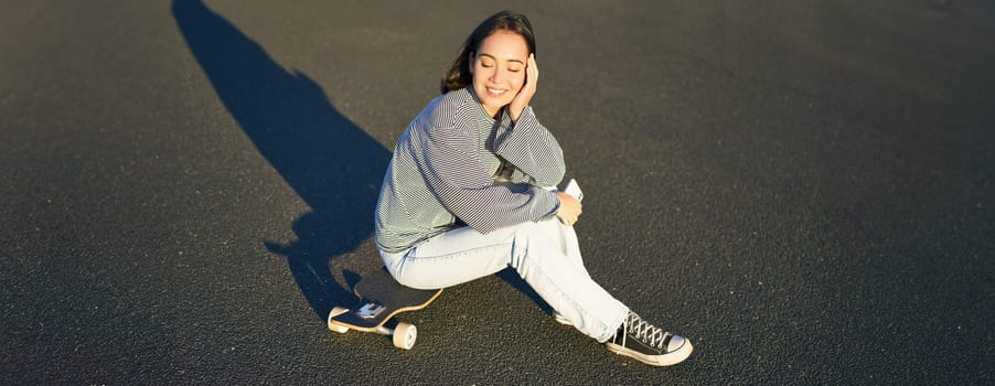 Portrait of asian woman sitting on skateboard, skating on her cruiser longboard, using smartphone app.