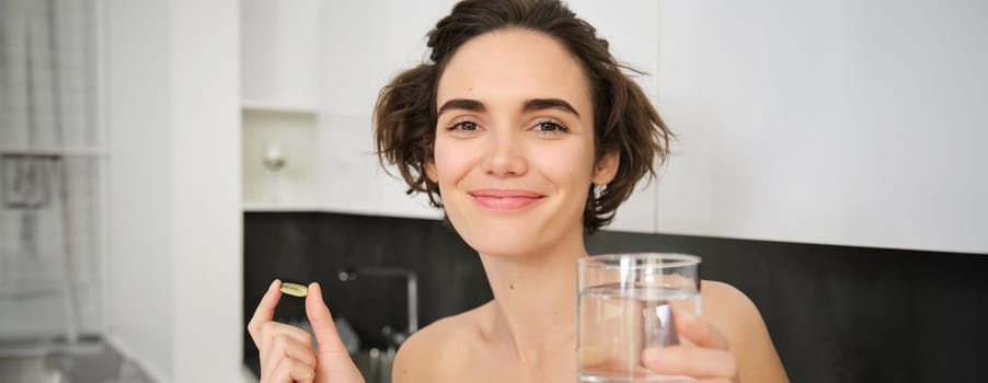 Dietry supplements and healthy lifestyle. Young woman taking vitamin C, D omega-3 with glass of water, standing in activewear, drinking after workout training in her kitchen.