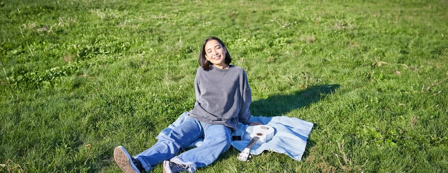 Young asian girl enjoying sunny day outdoors. Happy student having picnic on grass in park, playing ukulele.