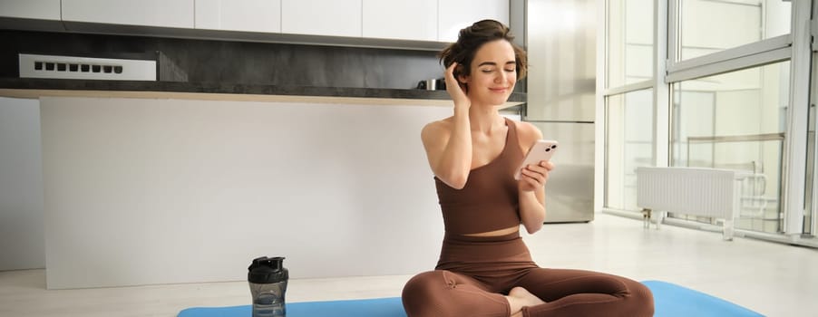 Sport and women lifestyle. Young woman does workout at home, sits on yoga mat with smartphone in bright kitchen, wears activewear, uses mobile phone app for indoor exercises.