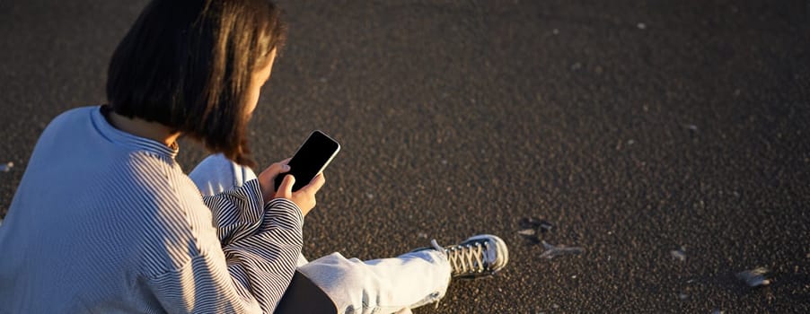 Vertical shot of asian girl typing, sending message or using mobile phone app, sitting on skateboard on road.